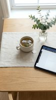 Cup of herbal tea on linen napkin beside a tablet and eucalyptus on a wooden table, minimal cozy workspace with natural light.