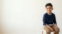Young boy with dark hair and olive complexion, wearing navy blue long-sleeve shirt and tan pants, sitting on wooden stool against neutral background. Studio portrait for advertising, education, and family-related projects.