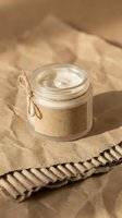 Open glass jar of white cream with kraft paper label and twine bow on crumpled brown paper background, natural organic skincare concept.
