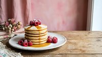 Stack of golden pancakes topped with fresh raspberries and butter on white plate, wooden table with pink floral background for breakfast menu design.