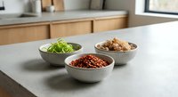 Three ceramic bowls with fresh herbs, spices and seasonings arranged on modern kitchen counter for culinary preparation and cooking ingredients display.