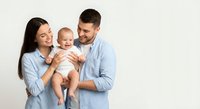 Happy young Caucasian family with infant baby in matching blue shirts smiling together against neutral background for parenting and lifestyle content.