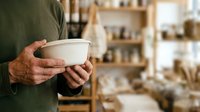 Close-up of male artisan hands holding empty ceramic bowl in pottery studio workshop with shelves of clay vessels and tools in background.