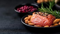 Raw salmon fillet with black pepper, almonds and rosemary sprig on black ceramic plate. Dark food photography with cranberries in background.
