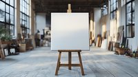 Empty white canvas on wooden easel in spacious art studio with natural lighting, large windows, and scattered art supplies. Professional creative workspace setting.