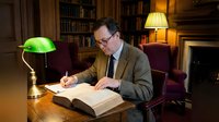 Middle-aged Caucasian businessman in suit writing in large book at wooden desk in traditional library office with green banker lamp and bookshelves.