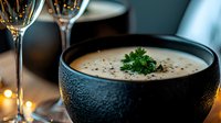 Creamy mushroom soup garnished with fresh parsley in black bowl, wine glasses and candlelight creating cozy restaurant atmosphere. Elegant dining setup.
