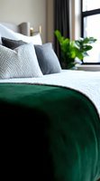 Cozy bedroom interior with emerald green velvet blanket, white textured bedding, gray pillows, and houseplants near window creating modern luxurious atmosphere.