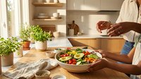 Person with medium skin tone preparing fresh vegetable salad in wooden bowl on kitchen counter with natural light, potted plants, and modern cabinetry for healthy eating and meal preparation content.