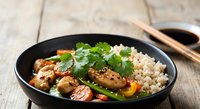 Asian stir-fry with grilled chicken, fresh vegetables and brown rice in black bowl on wooden table with chopsticks and soy sauce for healthy dining.