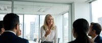 Confident blonde businesswoman presenting to diverse team in modern office conference room with city skyline view during corporate meeting.