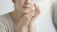 Young Asian woman with fair skin touching her face, wearing neutral beige t-shirt, expressing skincare, wellness, and beauty concerns in studio setting.