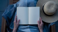 Young woman in denim shirt reading open blank book outdoors with straw hat, top view of hands holding empty journal for summer reading and relaxation.