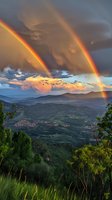 Dramatic double rainbow arches over mountain valley landscape at sunset, with stormy clouds and golden light illuminating rolling hills and distant village settlement.