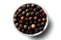 Aromatic allspice peppercorns in white ceramic bowl on white background, macro view of dried spice berries showing natural color variation and texture.
