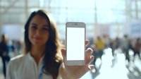 Young woman with long dark hair showing blank smartphone screen in crowded public space with soft focus background and natural lighting.