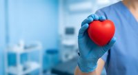 Healthcare professional in blue scrubs holding red heart symbol in gloved hands representing cardiology care and medical compassion in hospital setting.