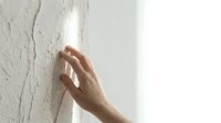 Hand touching cracked white wall surface showing structural damage and repair needs in residential interior space.