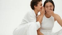 Two young women in white outfits share a secret, one whispering while the other laughs, captured on a light background.
