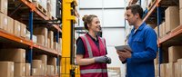 Two warehouse workers discussing inventory management in modern distribution center with organized shelving and safety equipment for logistics operations.