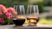Red and white wine glasses sitting outdoors on stone surface with pink flowering plants and blurred garden background, perfect for beverage, wine tasting, and lifestyle content.