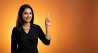 Smiling young Indian businesswoman in black blazer pointing upward with finger against orange gradient background for corporate presentations.