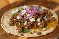 Ground meat taco with crumbled white cheese, purple onion rings and fresh cilantro on corn tortilla served on wooden cutting board, close up view.