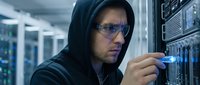 Male hacker in dark hoodie and glasses working with server equipment in data center, cybersecurity and network technology concept.