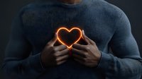 Young African American man in gray sweater holding glowing neon heart shape against chest on dark background, representing love, emotion and inner light concept.