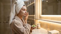 Young woman in bathrobe with towel on head applying skincare cream in modern bathroom mirror reflection spa wellness routine self-care morning ritual.
