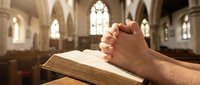 Hands clasped in prayer over open bible in gothic church interior with stained glass windows creating peaceful spiritual atmosphere for worship and meditation.