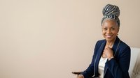 Mature African American female professional with gray hair in updo style wearing navy blazer and white top, smiling at camera while holding tablet device in modern office setting.