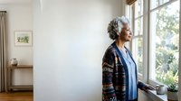 Senior woman with gray hair and light skin tone, wearing a dark patterned cardigan over blue clothing, gazing thoughtfully out a bright window in a minimalist home interior.