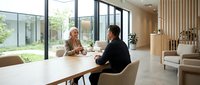 Young professional woman with light skin tone and middle-aged man with dark hair in business casual attire having discussion at modern white table in bright office space with large windows and contemporary furniture.