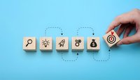 Business strategy concept with wooden blocks showing search, idea, growth, teamwork and money icons leading to target goal on blue background.