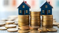 Small blue house models placed on stacks of gold coins against blurred background, representing real estate investment, property market and mortgage concepts.
