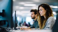 Young female professional with long wavy brown hair wearing gray sweater working focused at computer in modern office environment with coworkers in background.