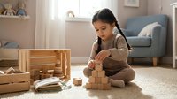 Young Asian girl in brown sweater playing with wooden building blocks on carpet in cozy living room with natural light and modern furniture.