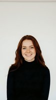 Happy young Caucasian woman with long red hair wearing black turtleneck sweater smiling at camera against neutral beige background for professional headshot.