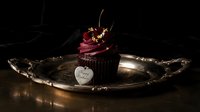 Chocolate birthday cupcake with dark berry frosting, cherry topping, gold flakes, and heart tag on vintage silver tray in moody dark setting.