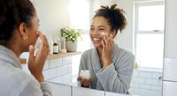 Young African American woman applying skincare cream in modern bathroom mirror, smiling during morning beauty routine with natural lighting and minimalist decor.