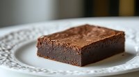 Rich, fudgy chocolate brownie square with dense texture and glossy chocolate frosting served on elegant white lace-patterned plate, close-up view of homemade dessert.