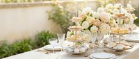 Elegant outdoor wedding reception table setting with white and pink floral centerpiece, crystal glasses, fine china plates and golden accents in garden.