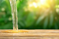 Clear stream of pure water pouring onto wooden surface against blurred green nature background with sunlight, representing freshness and purity.