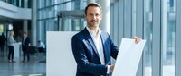 Confident middle-aged Caucasian businessman in navy suit presenting at flipchart in modern glass office building lobby during corporate meeting.