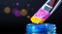 Vibrant paint brush dipped in pink and yellow paint dripping into blue liquid in glass jar, captured in macro on dark background with bokeh light effects.