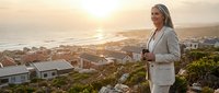 Mature woman with gray hair and glasses wearing light blazer stands on hillside overlooking coastal town at sunset, holding coffee cup, contemplating view for lifestyle and wellness content.