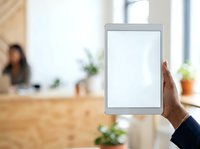 Close-up of male hand holding tablet with blank white screen in modern office workspace with blurred coworkers and plants in background.