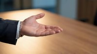 Caucasian male hand in open palm gesture wearing dark business suit and white dress shirt cuff on wooden table surface for corporate communication concepts.