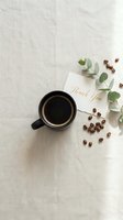Black coffee mug with thank you card, scattered roasted beans, and eucalyptus on linen. Flat lay styled scene for blogs and social media.